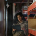 Karyawan gudang wanita mencatat stok barang dengan clipboard di antara rak penyimpanan